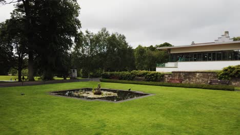 Dunfermline-glen-Fife-Scotland-with-lawn,-pond,-and-fountain-with-a-crow-stopping-to-drink-and-the-glen-pavilion-in-the-background