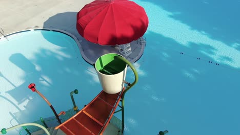 Rising-aerial-reveals-water-park-at-pool,-red-umbrellas-dot-a-sunny-day,-no-people