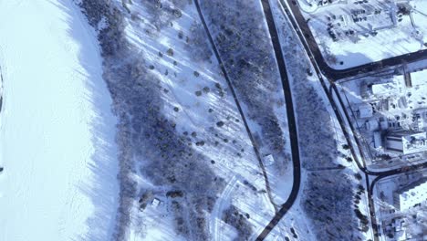 Aerial-birds-eye-view-over-winter-town-light-traffic-by-a-frozen-snow-cover-river-next-to-the-highway-by-a-town-community-residential-area-with-a-steap-road-leading-to-the-highway-freeway-forest-area
