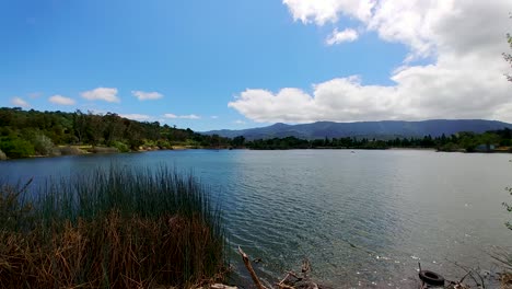 Lake-with-ripples-under-cloudy-blue-sky-surrounded-by-mountains-and-tall-grass