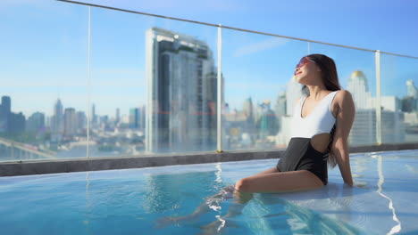 Beautiful-Asian-Woman-Sitting-in-Rooftop-Swimming-Pool,-City-Skyline