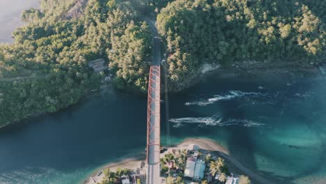 Aerial-View-Of-Steel-Bridge-Over-The-Turquoise-Blue-Water-With-Local-Fishing-Boats-Sailing-In-A-Small-Fishing-Village-In-Southern-Leyte,-Philippines---ascending-aerial-drone-shot,-pullback-shot