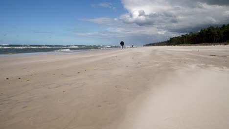 Windiger-Tag-An-Einem-Sandstrand,-Sandblasen,-Mann-Zu-Fuß-In-Der-Ferne