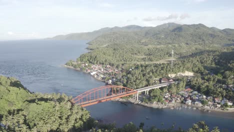 Overlooking-The-Blue-Water-Surrounded-By-Forest-Mountain-Under-The-Golden-Metal-Bridge-With-Vehicles-Travelling-Near-The-Small-Fishing-Village-In-A-Province-Of-Leyte,-Philippines