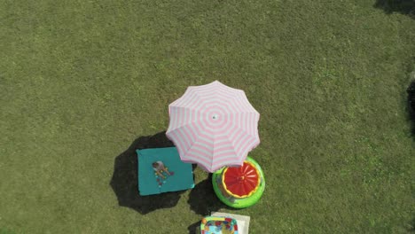 Top-Down-Aufnahme-Eines-Entzückenden-Kleinkindes,-Das-Auf-Der-Hellgrünen-Picknickmatte-Unter-Dem-Schönen-Rosa-Regenschirm-Im-üppigen-Garten-Sitzt---Aufsteigende-Drohne