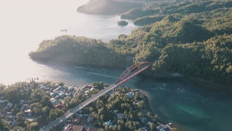 Vehicles-And-Motorcycles-Passing-By-The-Golden-Metal-Bridge-Connecting-The-Small-Fishing-Village-And-The-Lush-Mountains-Overlooking-The-Tropical-Ocean-In-Leyte,-Philippines