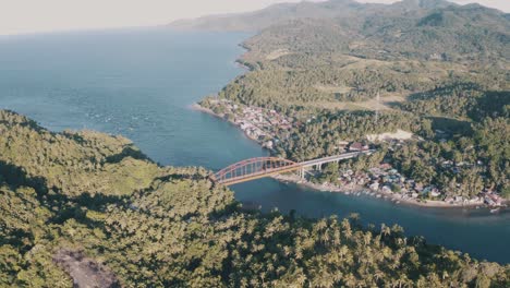 Puente-De-Metal-Dorado-Que-Se-Extiende-Sobre-Las-Tranquilas-Aguas-Azules-Que-Conectan-El-Pequeño-Pueblo-De-Pescadores-Y-Las-Exuberantes-Montañas-Con-Verdes-Cocoteros-En-El-Sur-De-Leyte,-Filipinas