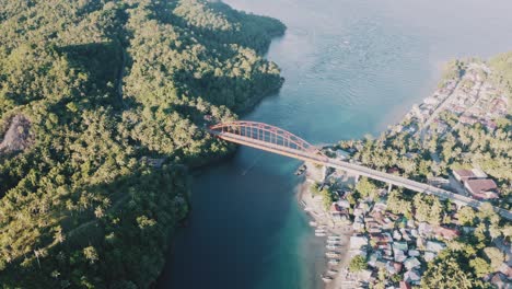 Barco-De-Pesca-Navegando-En-El-Agua-Azul-Bajo-El-Puente-De-Metal-Dorado-Que-Conecta-Las-Exuberantes-Montañas-Y-El-Pequeño-Pueblo-De-Pescadores-En-Una-Provincia-En-El-Sur-De-Leyte,-Filipinas
