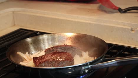 Steaks-and-onions-cooking-in-a-stainless-steel-frying-pan-on-a-gas-stove