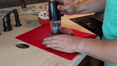 Feminine-hands-seasoning-raw-steak-with-salt-and-pepper-in-a-small-kitchen