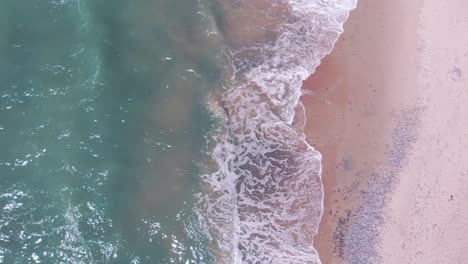 Top-aerial-view-of-ocean-waves-splash-against-pink-tropical-beach-on-summer-day,-zooming-out