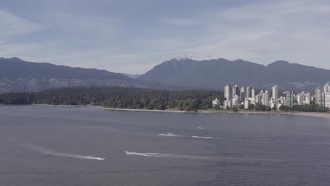 Aerial-fly-over-Kitsilano-Beach-to-English-Bay-Stanley-Park-as-leisure-motor-boats-zip-by-sail-boats-parked-people-suntanning-fishing,-hugging-and-romantic-relaxation-on-a-hot-summer-day-in-BC-1-4