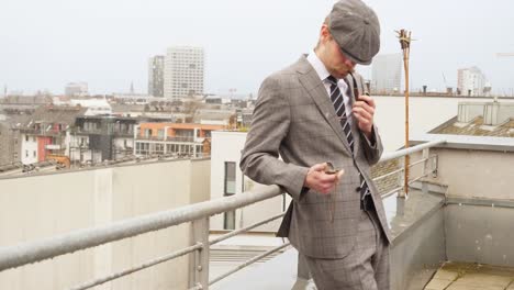 a-well-dressed-man-with-a-beret-looking-on-his-pocket-watch-while-smoking-an-oldschool-pipe-on-the-rooftop-during-a-foggy-wintersday