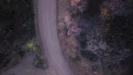 Dark-Vehicle-Drives-Down-Curved-Dirt-Road-Through-Dense-Forest-at-Dusk