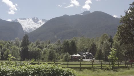 Summer-Mountainside-snow-covered-below-with-a-ranch-ofr-livestock-and-stallions-with-a-luxury-home-on-a-hill-with-wooden-fencing-backdrop-of-lush-green-forest-coverage-with-a-gleam-of-fantastic-beauty