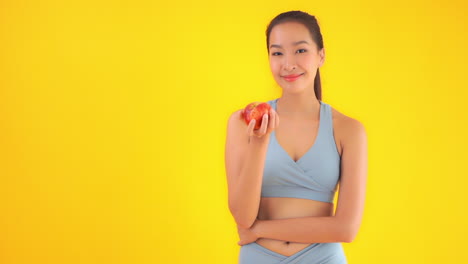 Asiatische-Frau-In-Sportbekleidung-Im-Fitnessstudio-Mit-Frischem-Rotem-Apfel,-Zeitlupe,-Gesundem-Essen-Und-Lifestyle-Konzept,-Statische-Aufnahme-Isoliert-Auf-Gelbem-Hintergrund