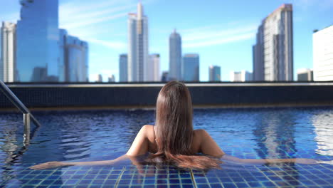 Back-of-Classy-Female-With-Long-Hair-Enjoying-in-Rooftop-Pool-With-Stunning-View-on-Modern-City-and-Skyscrapers-of-Bangkok,-Full-Frame