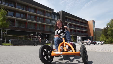 Rotating-FHD-shot-of-a-small-girl-riding-an-orange-pedal-quadricycle-and-zigzagging-on-an-asphalt-road-in-a-private-resort-in-Dolní-Morava,-Czech-Republic