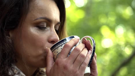Close-up-FHD-shot-of-a-brunette-middle-aged-woman-with-her-eyes-closed-savoring-a-cup-of-fresh-hot-tea-in-the-morning-with-trees-in-the-background