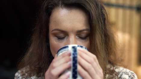 Frontal-close-up-FHD-shot-of-a-woman-holding-a-cup-with-both-hands-and-drinking-freshly-poured-hot-tea,-savoring-the-taste-with-a-wooden-house-in-the-background