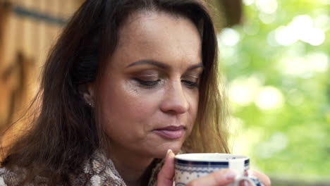 Close-up-FHD-shot-of-a-middle-aged-brunette-woman-enjoying-the-smell-of-a-fresh-cup-of-hot-tea-for-breakfast-in-front-of-a-weekend-house-in-nature