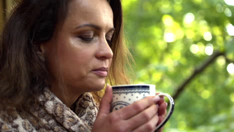 Close-up-FHD-shot-of-a-brunette-middle-aged-woman-smelling-freshly-poured-tea-in-a-mug-she-holds-with-both-her-hands-with-blurred-trees-in-the-background