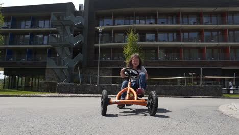 Rotating-FHD-shot-of-a-small-girl-cycling-on-an-orange-pedal-quadricycle-in-a-private-resort-in-Dolní-Morava,-Czechia-with-her-family-cycling-behind