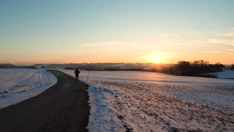 Eine-Person-Läuft-Eine-Einsame-Straße-Entlang,-Die-Sich-In-Einer-Wunderschönen-österreichischen-Winterlandschaft-Befindet,-Während-Die-Sonne-Untergeht