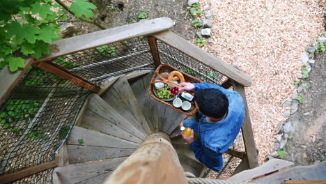 Static-overhead-FHD-shot-of-a-man-in-a-denim-jacket-and-jeans-climbing-a-spiral-wooden-staircase-carrying-a-basket-with-breakfast