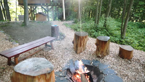Tilt-FHD-shot-of-a-burning-campsite-fire-surrounded-by-stumps-for-sitting-and-a-private-luxurious-treehouse-accommodation-behind-in-Dolní-Morava,-Czechia