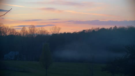 A-beautiful-vivid-early-spring-sunset-on-the-country-side,-featuring-a-house-releasing-smoke-in-the-background,-and-trees-in-the-mid-ground-and-foreground