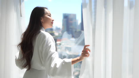 Pretty-young-asian-woman-moving-curtains-from-windows-on-sunny-morning,-slowmo