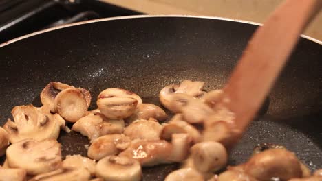 Close-up-of-sliced-button-mushrooms-sauteing-in-butter,-stirred-with-wooden-spoon