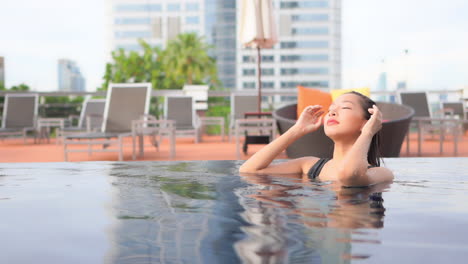 Beautiful-asian-female-fixing-her-hair-after-swimming-in-a-luxury-pool-with-city-in-background,-full-frame-slow-motion