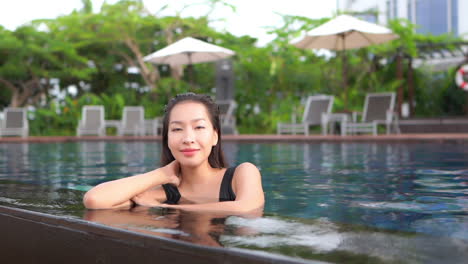 While-leaning-on-the-edge-of-a-resort-swimming-pool,-a-pretty-young-woman-cocks-her-head-and-smiles