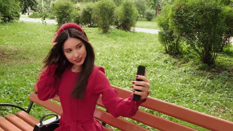 Pretty-young-brunette-in-red-sits-on-a-park-bench-and-takes-a-selfie