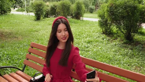 Beautiful-girl-in-red-sits-on-a-park-bench-and-takes-a-selfie