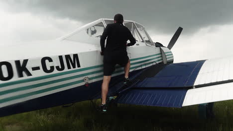 Following-FHD-shot-of-a-man-climbing-on-a-wing-of-a-small-sport-airplane,-preparing-to-enter-the-cabin-and-take-off-from-a-field-airport-with-a-grey-stormy-sky-above
