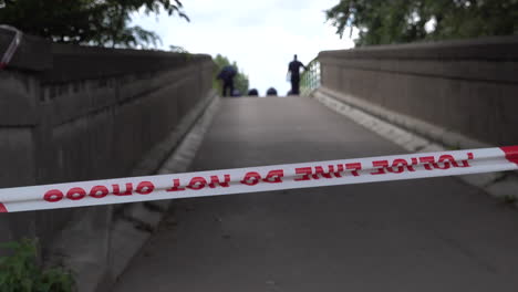 A-London-Metropolitan-police-forensics-team-working-on-their-hands-and-knees-to-investigate-and-search-a-bridge-behind-red-police-cordon-tape,-following-a-gun-and-knife-crime-incident