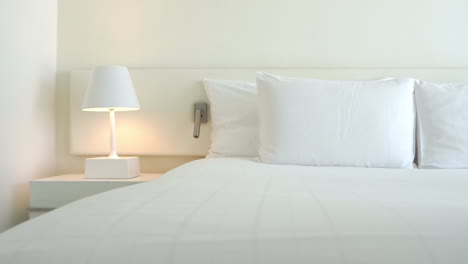 In-a-white-hotel-room,-there-is-a-white-lamp-on-a-white-bedstand-illuminating-a-headboard-and-the-crisp-white-linen-of-a-perfectly-made-up-bed