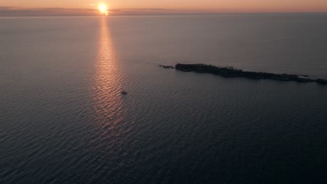 Solitary-Sailboat-Sailing-Through-The-Sea-Near-An-Island-During-Sunset---aerial-shot