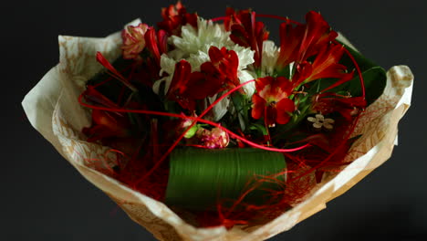 Zoomed-out-a-large-bouquet-of-red-and-white-flowers-and-green-decorative-elements-in-a-glass-vase-decorated-with-paper-around-on-a-dark-background