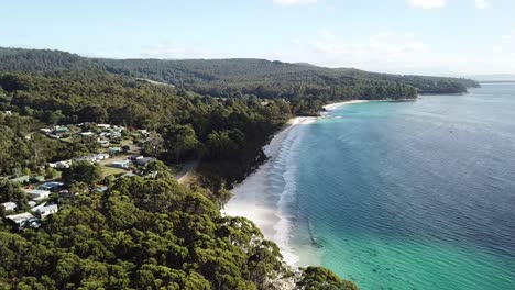 Aerial-drone-pan-over-stunning-isolated-beach-and-coastal-town-on-sunny-day-with-aqua-blue-water-and-white-sand-surrounded-by-green-forest-as-waves-roll-in