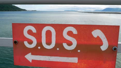 Painted-red-SOS-sign-handing-on-promenade-railings-ocean-safety-with-coastline-horizon-push-in
