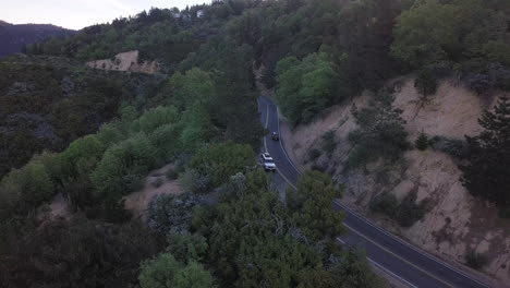 Dark-SUV-truck-drives-around-a-curve-in-a-two-lane-highway-through-a-mountain-pine-forest-at-dusk
