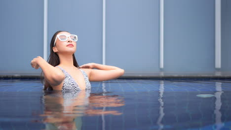 Young-attractive-asian-female-fixing-her-wet-hair-in-swimming-pool,-hairstyling-and-haircare-products-concept,-full-frame-slow-motion-with-copy-space