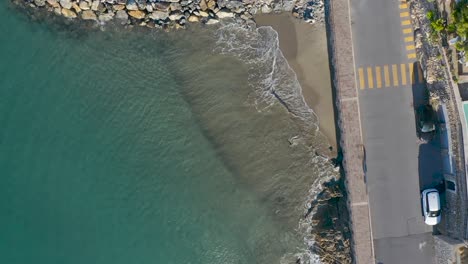 top-down-drone-shot-of-a-beach-near-a-road-in-Liguria,-slowly-tilting-up-and-revealing-the-village-of-Alassio