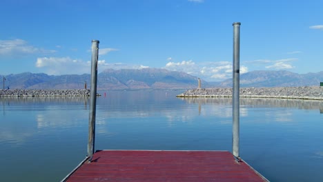 Fixed-shot-from-the-end-of-a-red-wooden-jetty-looking-out-towards-the-Saratoga-Springs-Marina-entrance-with-flat-water-and-crystal-clear-mountains-in-the-background,-only-a-few-clouds-in-the-distance