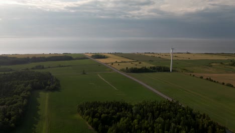 Wind-Turbines-Spinning-On-The-Lush-Fields-In-Quebec,-Canada-With-Cars-Driving-On-The-Asphalt-Road-On-A-Cloudy-Day---aerial-drone,-panning-shot