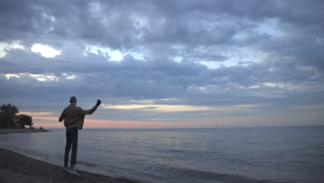 Mann-Springt-Und-Streckt-Sich-Fröhlich-Am-Strand-Mit-Blick-Auf-Einen-Wunderschönen-Rosa-Und-Violetten-Sonnenuntergang---Weitschuss